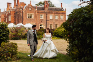 Bride and groom walk hand in hand across the lawn at Hodsock Priory, the Tudor house and marquee behind them