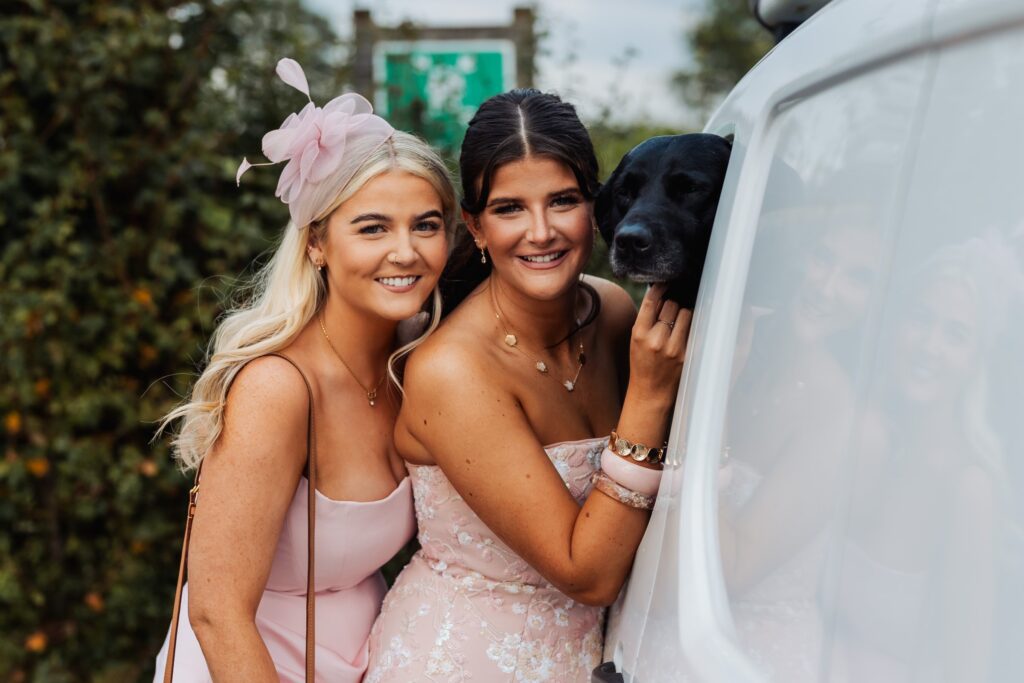 Two guests posing beside a car with a black dog looking out of the window