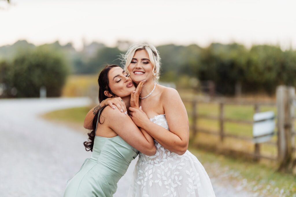 Bridesmaid hugging the bride cheek to cheek in the countryside at Sedgewell Barn