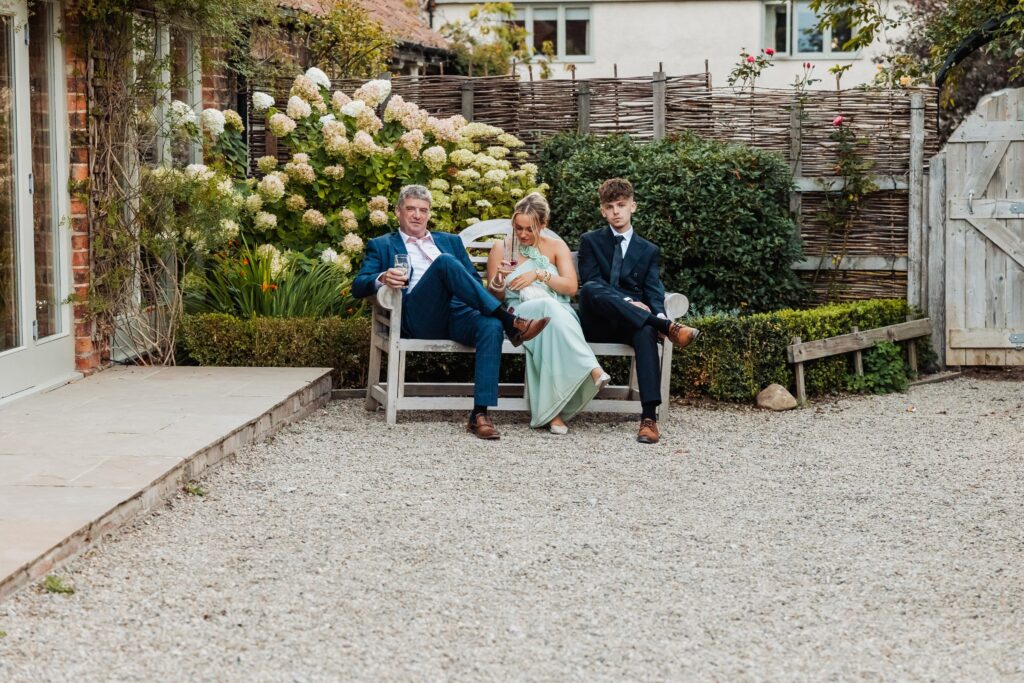 Wedding guests sitting on a bench beside hydrangeas outside Sedgewell Barn