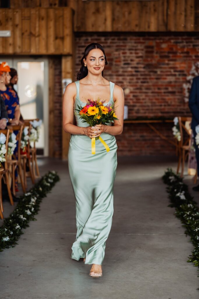 Bridesmaid walking down the aisle carrying an orange and sunflower bouquet