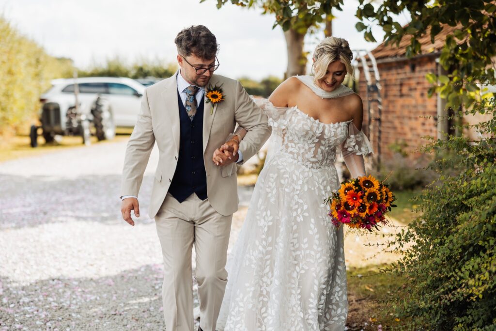 Bride and groom walking up the drive at Sedgewell Barn with a sunflower bouquet