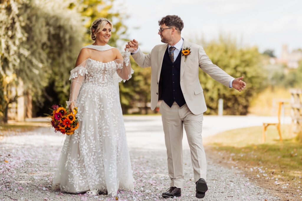Bride and groom walking hand in hand at Sedgewell Barn, the bride carrying a bright sunflower bouquet