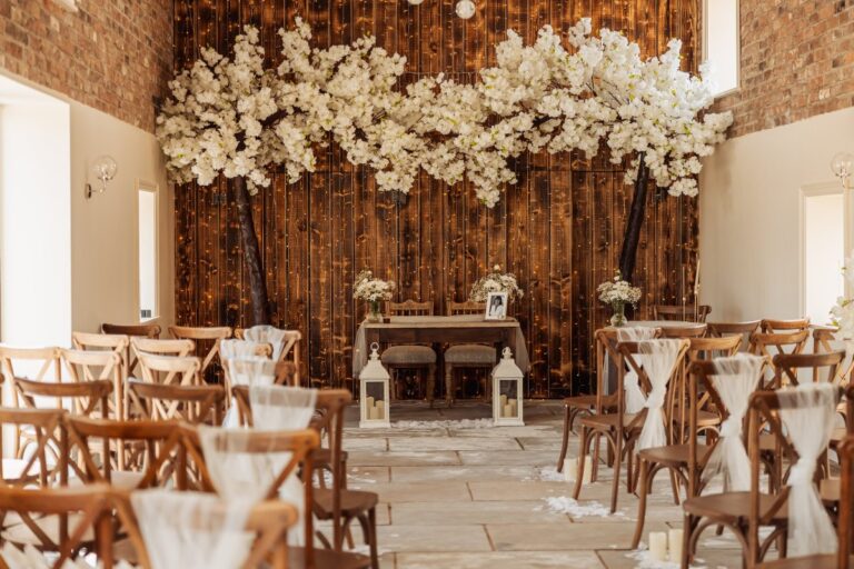 Dove Barn set for the ceremony at Clayfield Farm, with a white blossom canopy arching over the registrar's table and cross-back chairs dressed in chiffon.