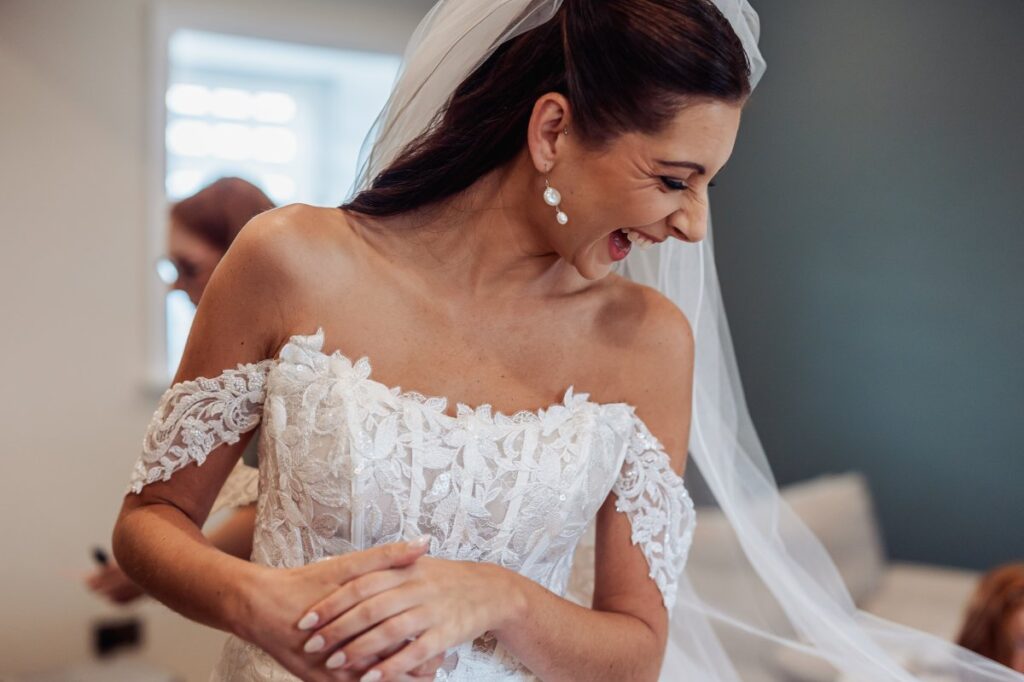 The bride laughs during bridal preparations, wearing an off-shoulder lace gown and a full-length veil.