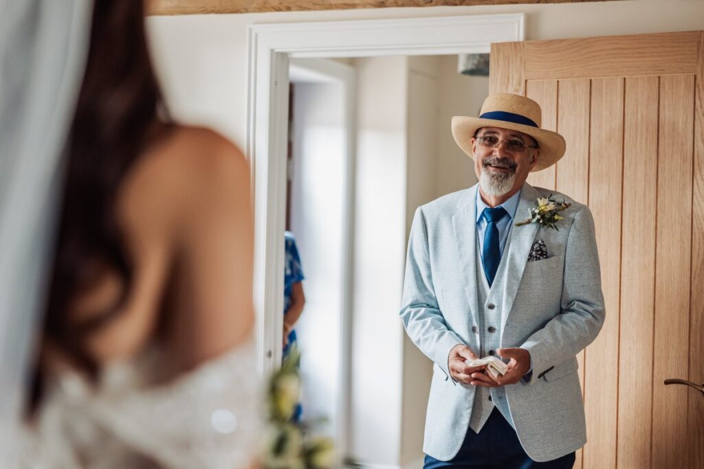 The bride's father, in a straw boater and pale grey suit, sees his daughter in her wedding dress for the first time.