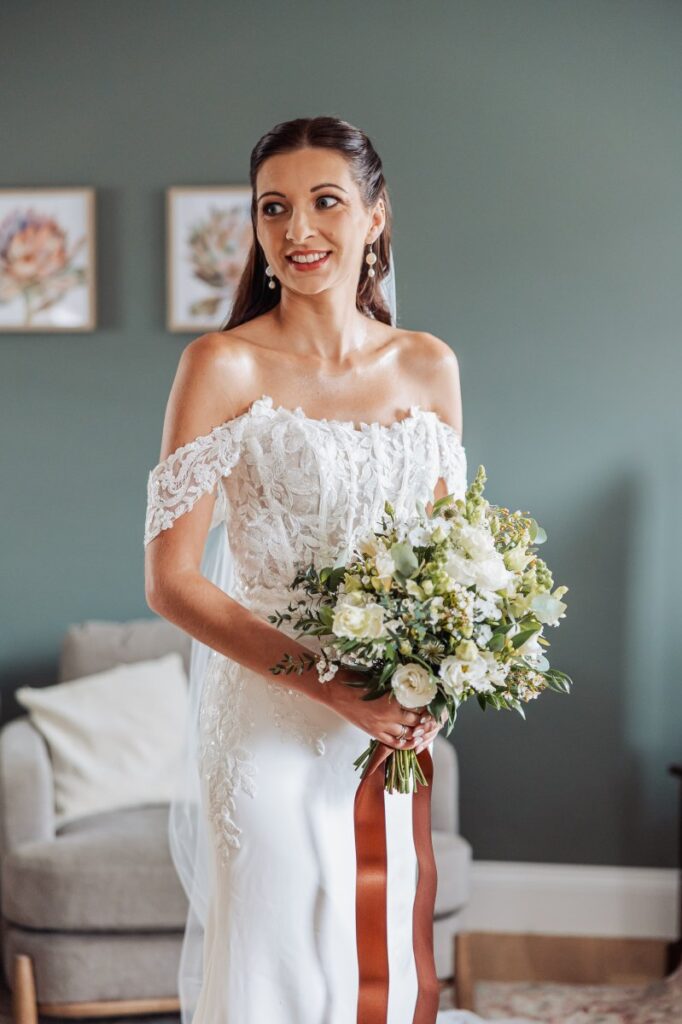 Bridal portrait against a sage green wall, holding a white and green bouquet finished with trailing rust-brown ribbon.