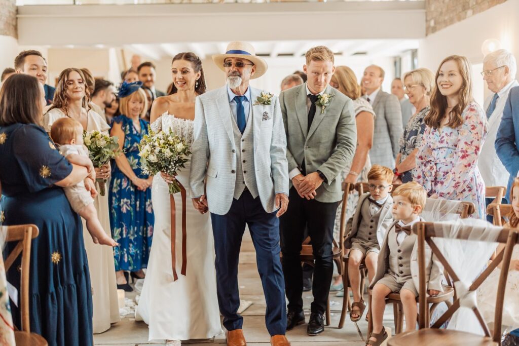 The bride walks down the aisle on her father's arm as guests stand in the light-filled ceremony barn.
