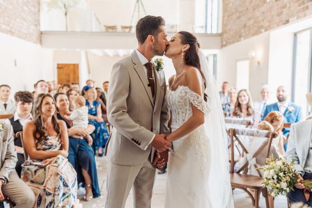 The first kiss as husband and wife, watched by seated guests beneath the white beams of Clayfield Farm's ceremony barn.