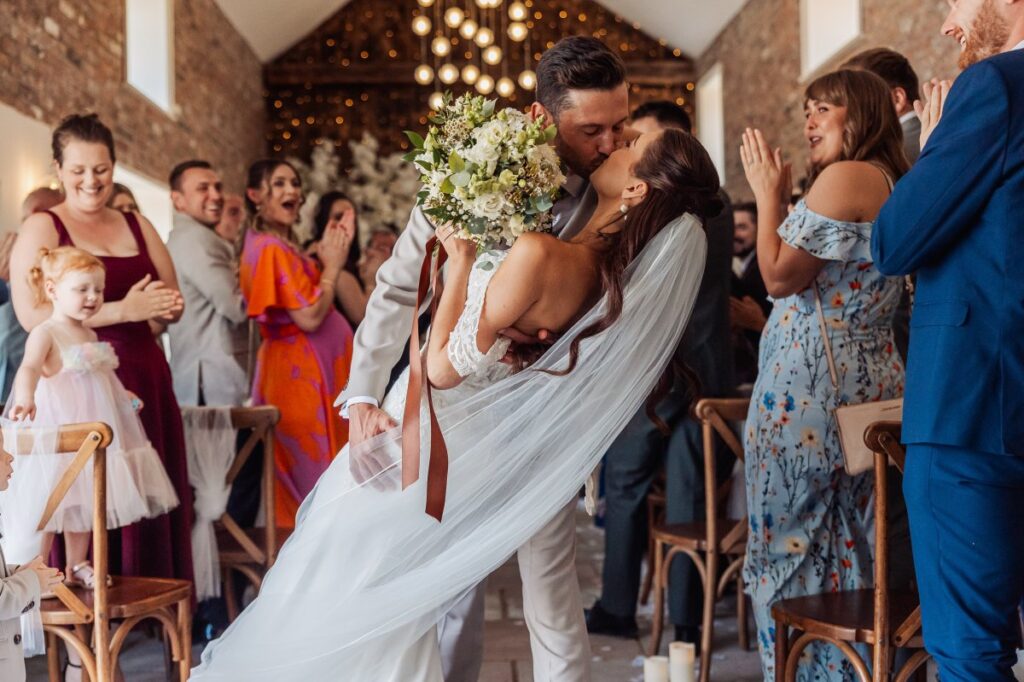 The groom dips his bride for a kiss as guests applaud, bouquet raised, under the fairy-lit rafters at Clayfield Farm.