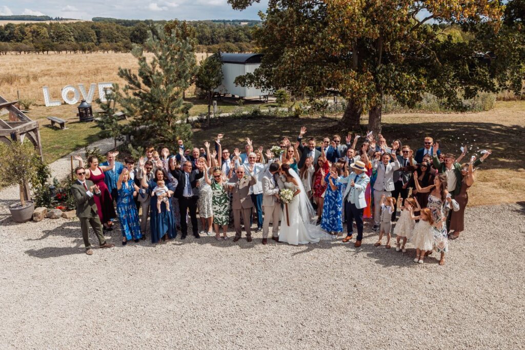 The full wedding party gathered on the gravel at Clayfield Farm, with the Yorkshire Wolds and a shepherd's hut behind them.