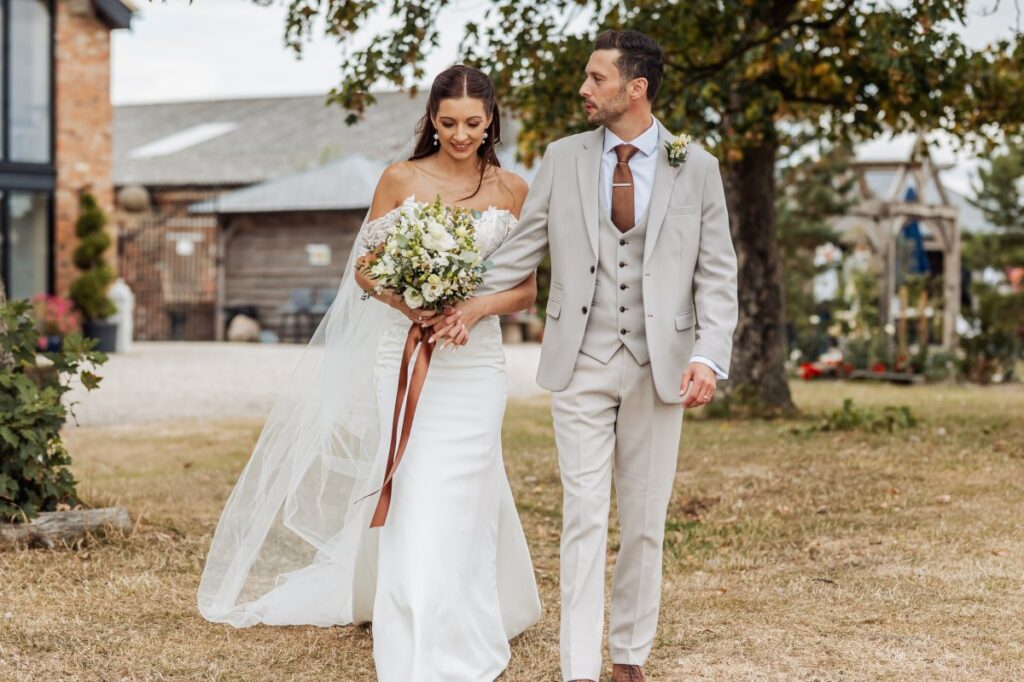 Bride and groom walk together past the brick barns at Clayfield Farm, her veil trailing across the grass.