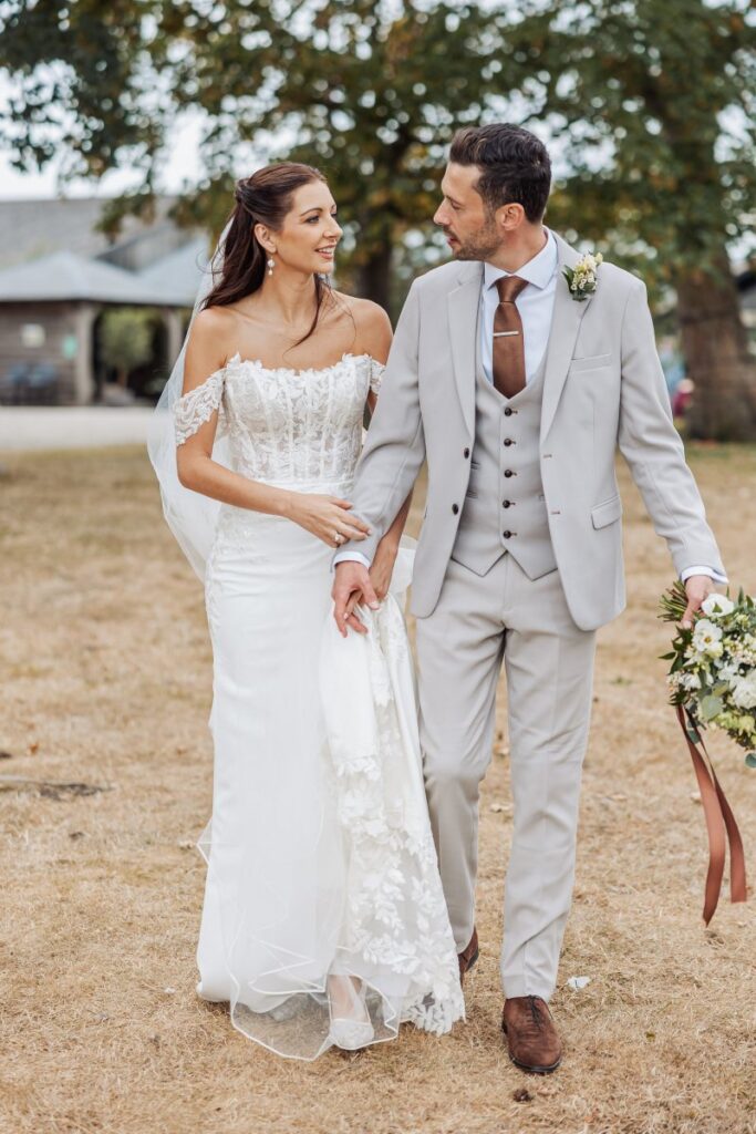The couple walk through the sun-dried grass at Clayfield Farm, the groom carrying her bouquet.