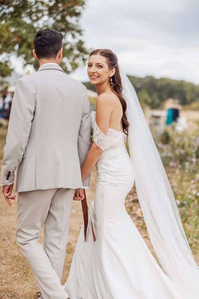The bride glances back over her shoulder, her veil running the full length of her fitted lace gown.