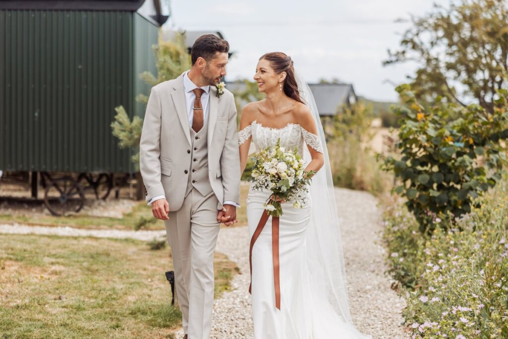 Bride and groom walk the gravel path past the shepherd's huts at Clayfield Farm.