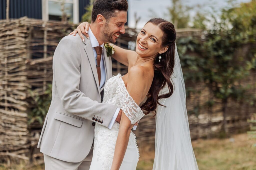 The bride smiles over her shoulder as her husband holds her, against the woven willow fence at Clayfield Farm.