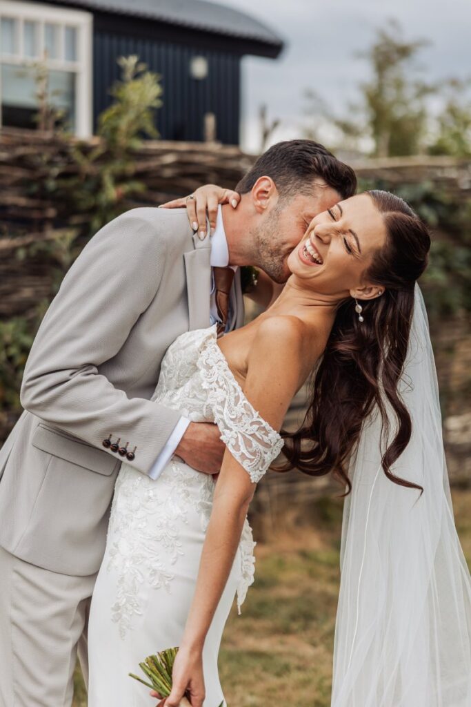 The groom kisses his bride's cheek as she laughs, photographed in the gardens at Clayfield Farm.