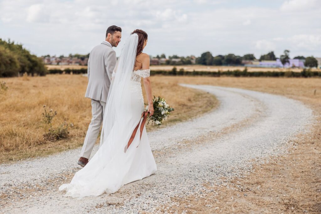 Bride and groom walk the farm track at Clayfield Farm, the dry summer fields of the Yorkshire Wolds stretching behind them.