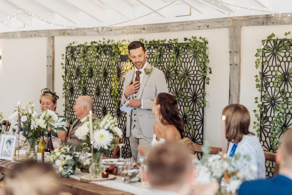 The groom delivers his speech in the oak-framed reception room at Clayfield Farm, backed by foliage screens.
