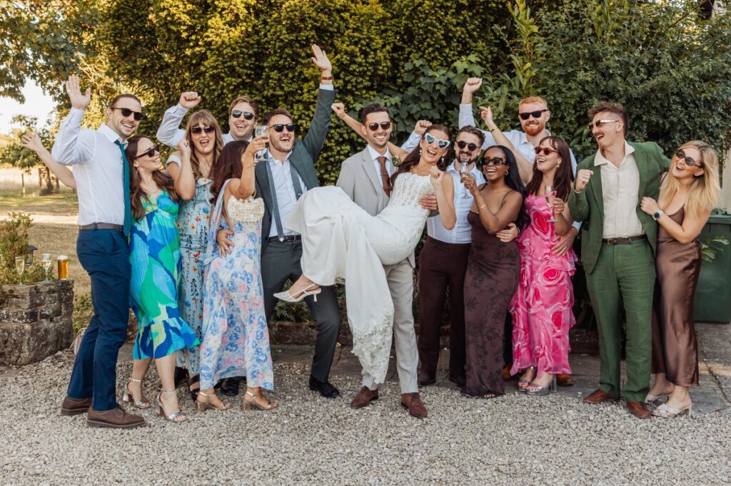 Friends cheer as the groom lifts his bride for a group photograph in the evening sun.