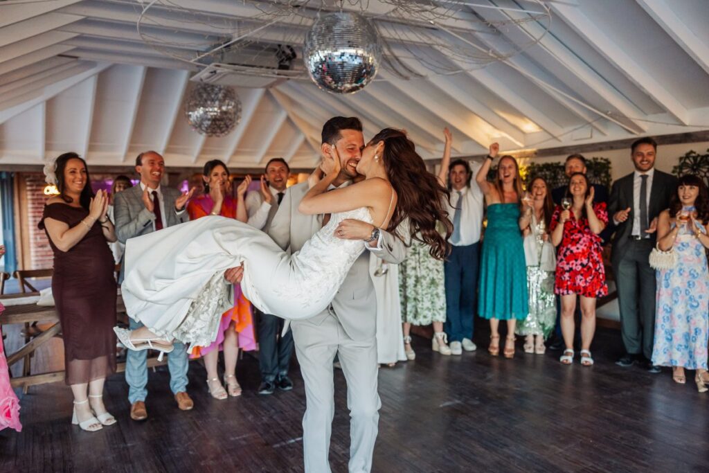 The groom lifts his bride during their first dance as guests cheer beneath the disco balls at Clayfield Farm.
