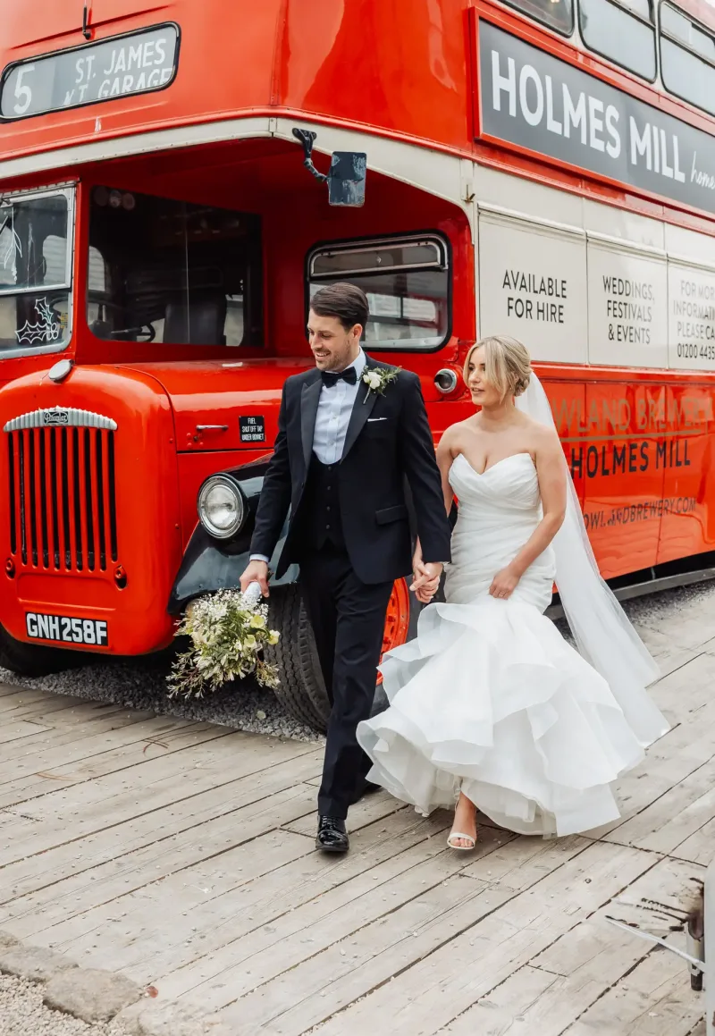 A bride and groom walk hand in hand in front of a vintage red double-decker bus with Holmes Mill signage in Clitheroe. The groom wears a black tuxedo; the bride, in a white strapless gown, holds her train, capturing the charm of their wedding day.