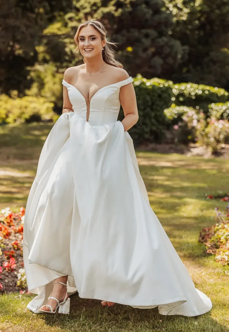A smiling bride in an off-the-shoulder white wedding gown holds her dress while walking through a sunny garden at Whirlow Brook Hall, with colorful flowers and greenery in the background.